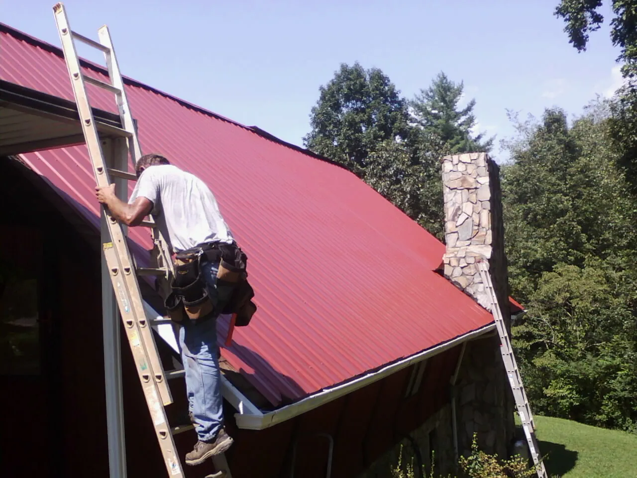 Roofer on a ladder beside a red metal roof and stone chimney — Soffit & Fascia Repair by Roofing Brothers in Glenoma